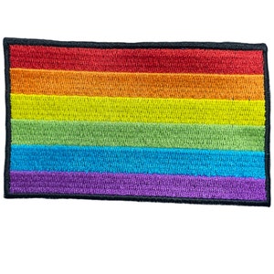 Op de afbeelding: Een regenboog-pride-vlagpatch met zes horizontale strepen in de kleuren rood, oranje, geel, groen, blauw en paars. De patch is omgeven door een zwarte rand.