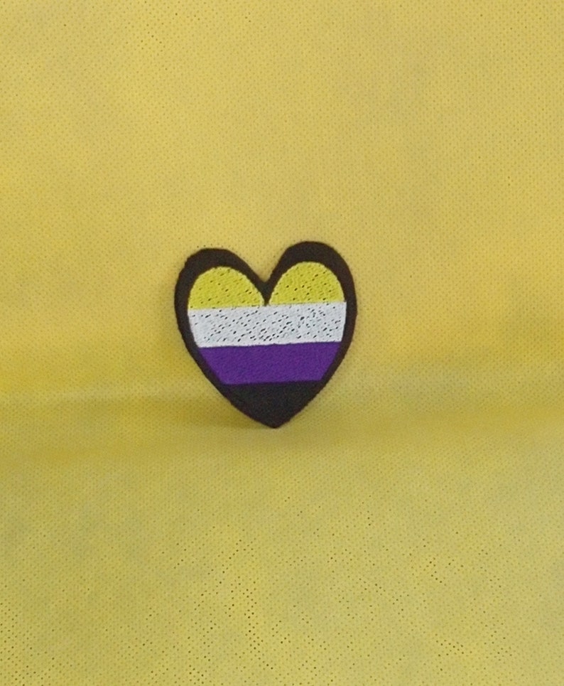 May include: A yellow, white, purple, and black heart-shaped patch with a stitched design. The patch is on a yellow background.