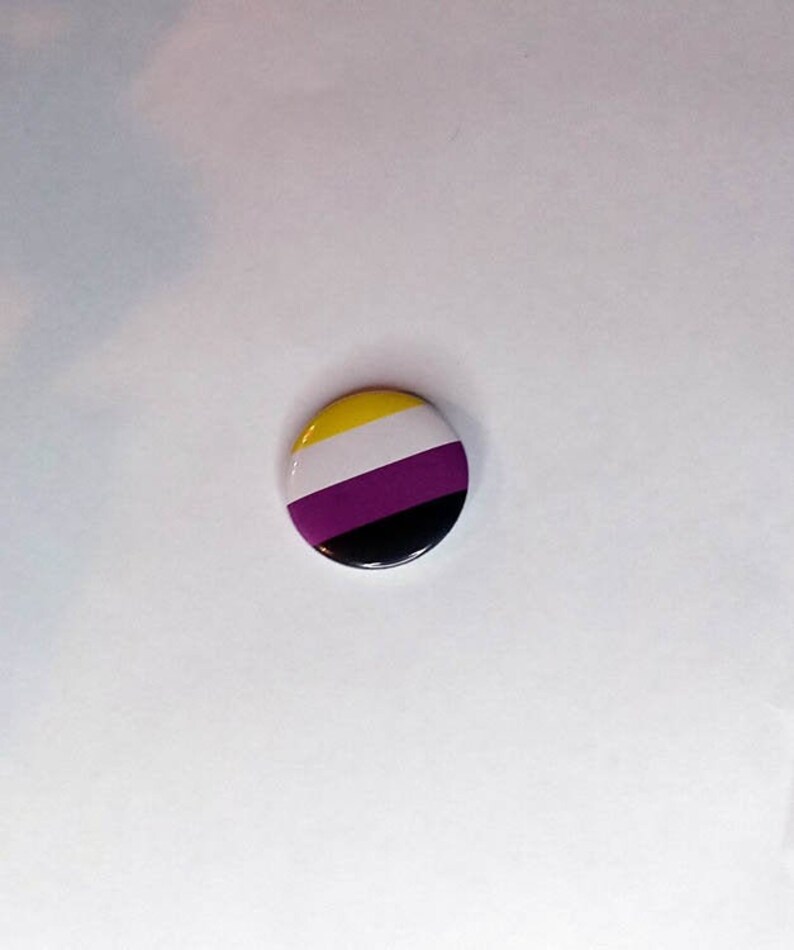 以下が含まれることがあります： 黄色、白、紫、黒のストライプ模様の丸いボタン。このボタンはノンバイナリーアイデンティティの象徴です。