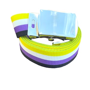 Puede incluir: Un cinturón con los colores del orgullo no binario, con un patrón de rayas amarillas, blancas, moradas y negras. El cinturón tiene una hebilla plateada brillante. Hecho de tela tejida.