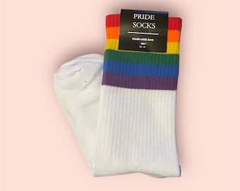 May include: White socks with a rainbow stripe pattern. The socks have a black tag that says "PRIDE SOCKS" and "made with love".