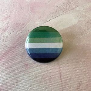 Op de afbeelding: Een ronde knop met een groen, wit, blauw en zwart gestreept ontwerp. De knop is een representatie van de genderqueer pride-vlag.