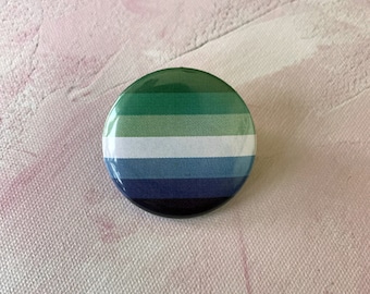 Puede incluir: Un botón redondo con un diseño a rayas verde, blanco, azul y negro. El botón es una representación de la bandera del orgullo genderqueer.