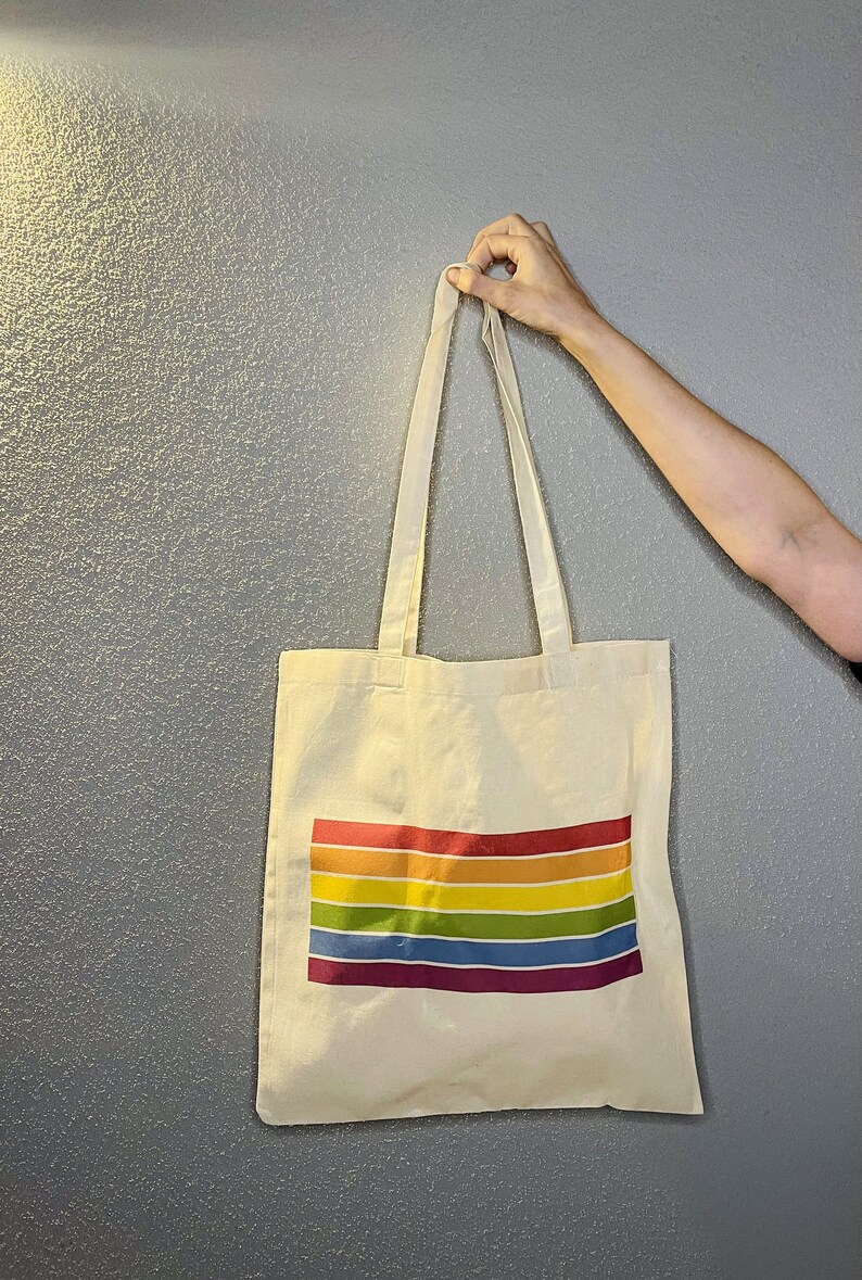 May include: A white canvas tote bag with rainbow stripes printed on the front. The bag has two long straps.