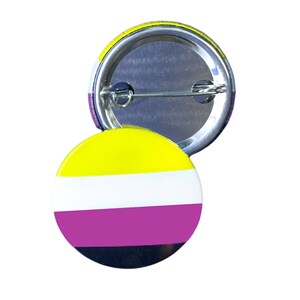 以下が含まれることがあります： ノンバイナリープライドフラッグの色をした丸いボタン。ボタンは黄色、白、紫、黒です。銀色のピンが付いています。