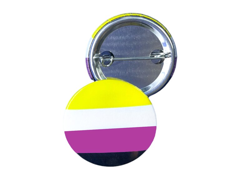 以下が含まれることがあります： ノンバイナリープライドフラッグの色をした丸いボタン。ボタンは黄色、白、紫、黒です。銀色のピンが付いています。