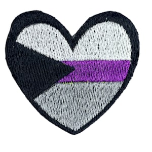 Puede incluir: Un parche bordado en forma de corazón con los colores de la bandera del orgullo asexual: negro, blanco, gris y morado.