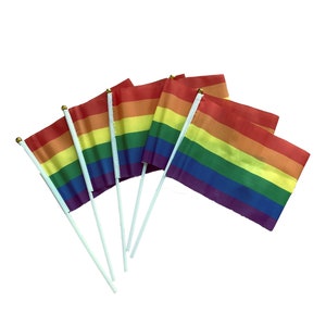 Op de afbeelding: Een set van vijf regenboog-pride-vlaggen op witte stokken. De vlaggen zijn in een waaier vorm gerangschikt, met de rode strepen naar buiten en de paarse strepen naar binnen.