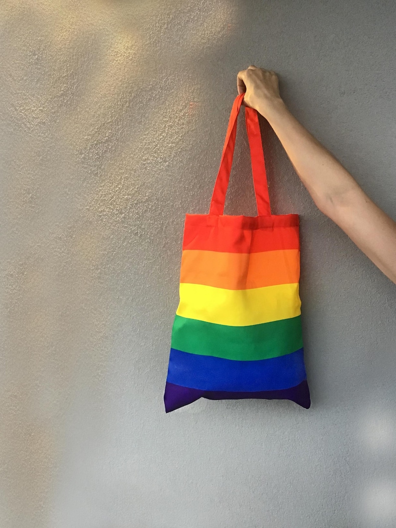 May include: A rainbow tote bag with red, orange, yellow, green, blue, and purple stripes. The bag is hanging from a hand.