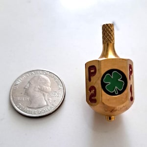 May include: A gold-colored metal dreidel featuring a green four-leaf clover emblem on a black circular background, with the red letters P and 2 visible on the side, positioned next to a United States quarter coin for scale reference.