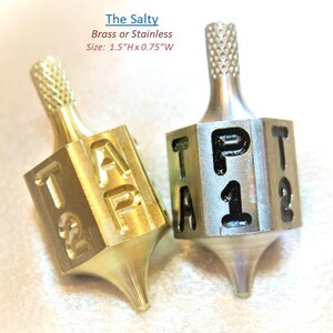 May include: Two metal spinning tops, one brass and one stainless steel, with the text "TAP 12" engraved on the sides. The brass top is gold-colored, and the stainless steel top is silver. Each top is approximately 1.5 inches high and 0.75 inches wide.