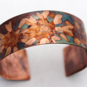 May include: A copper cuff bracelet with a hammered finish and a swirling, abstract pattern in shades of brown, copper, and blue.