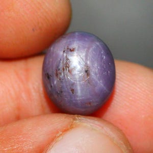 Op de afbeelding: Een paarse ster-saffier edelsteen met een zespuntige ster in het midden. De edelsteen is cabochon geslepen en heeft een glad, afgerond oppervlak.