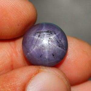 Op de afbeelding: Een ronde, lavendelkleurige edelsteen met een stervormig patroon. De edelsteen wordt tussen vingers gehouden, wat het gepolijste oppervlak en de natuurlijke insluitsels benadrukt. De kleur van de steen verandert in het licht, wat een boeiend visueel effect creëert.