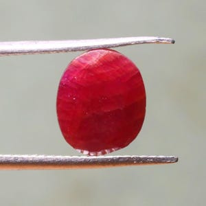 Puede incluir: Una gema ovalada de color rojo vibrante sostenida por unas pinzas. La gema tiene una superficie lisa y pulida y una apariencia ligeramente transl&uacute;cida.