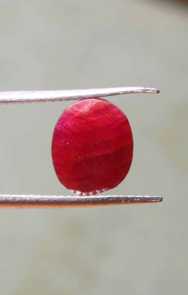Puede incluir: Una gema ovalada de color rojo vibrante sostenida por unas pinzas. La gema tiene una superficie lisa y pulida y una apariencia ligeramente transl&uacute;cida.