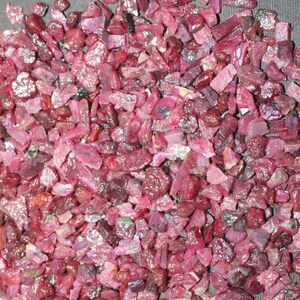 May include: A pile of raw, uncut ruby gemstones. The stones vary in size and shape, displaying a range of pink and deep red hues. The rough texture and natural forms of the rubies are visible.