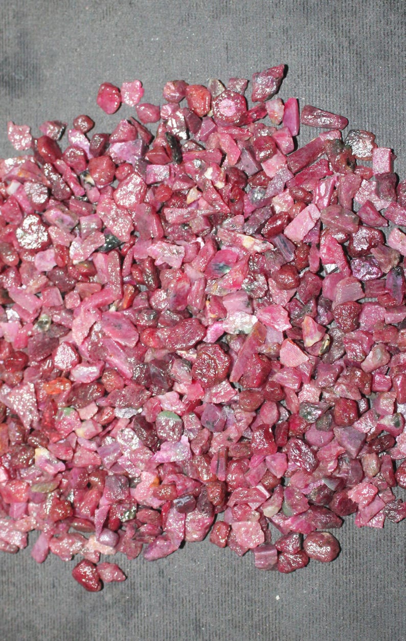 May include: A pile of raw, uncut ruby gemstones. The stones vary in size and shape, displaying a range of pink and deep red hues. The rough texture and natural forms of the rubies are visible.