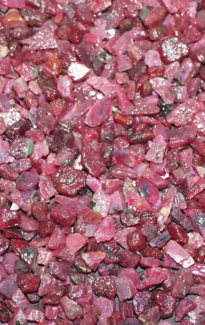 May include: Close-up of a pile of rough-cut ruby gemstones. The stones vary in size and shape, displaying a range of deep red hues with some lighter pink and purple tones. The surface of the stones appears slightly reflective.
