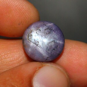 Puede incluir: Una piedra preciosa redonda de color púrpura con un patrón en forma de estrella en el centro. La piedra preciosa se sostiene en una mano.