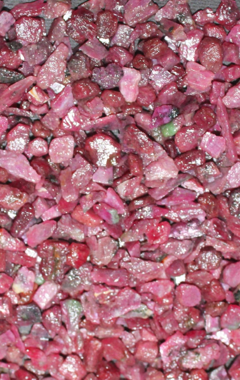 May include: A close-up view of a pile of small, rough, red ruby gemstones. The rubies are various shapes and sizes, and they have a shiny, polished finish.