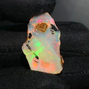 May include: A raw opal gemstone with a vibrant display of colors. The opal features a milky white base with flashes of green, pink, and orange. Dark inclusions and a brown patch add contrast. The gemstone is held against a black background.