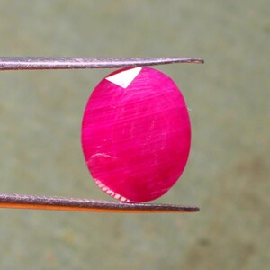 K&ouml;nnte beinhalten: Ein leuchtend rosa ovaler Edelstein, der mit einer Pinzette gehalten wird. Der Edelstein hat eine glatte, polierte Oberfl&auml;che und einen leichten Schimmer.