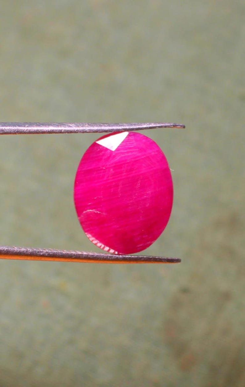 K&ouml;nnte beinhalten: Ein leuchtend rosa ovaler Edelstein, der mit einer Pinzette gehalten wird. Der Edelstein hat eine glatte, polierte Oberfl&auml;che und einen leichten Schimmer.