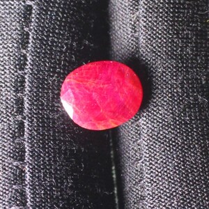 Puede incluir: Una gema ovalada de color rojo brillante con un corte facetado. La gema est&aacute; colocada sobre un fondo de tela negra.