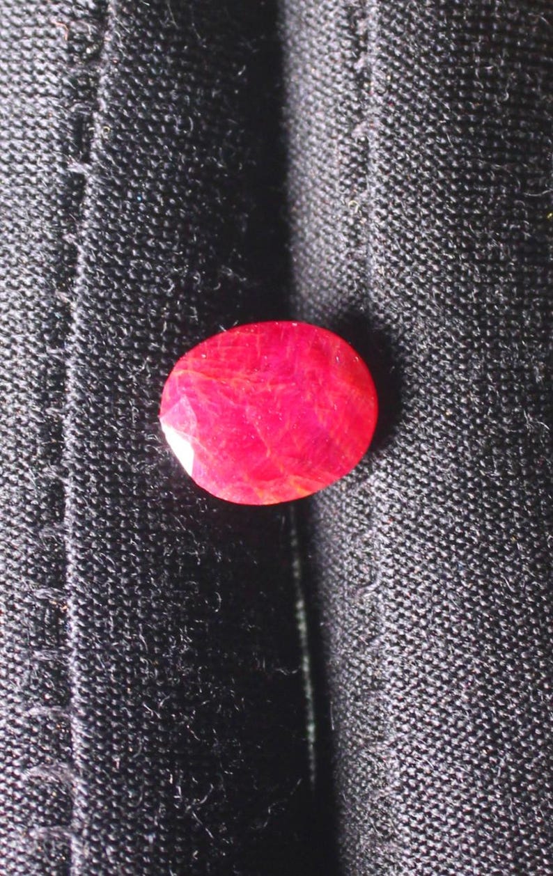 Puede incluir: Una gema ovalada de color rojo brillante con un corte facetado. La gema est&aacute; colocada sobre un fondo de tela negra.