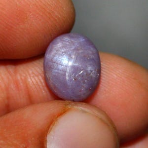 Puede incluir: Primer plano de una gema ovalada de color lavanda. La gema tiene una superficie lisa y pulida con sutiles líneas internas y un efecto de estrella. La piedra se sostiene entre dos dedos sobre un fondo borroso.