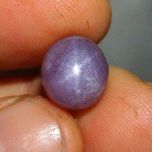 Op de afbeelding: Een ronde, gepolijste, lavendelkleurige edelsteen die tussen vingers wordt gehouden. De edelsteen heeft een stervormig effect op het oppervlak, met een centraal punt en uitstralende lijnen. De kleur van de steen is een zacht, gedempt paars.