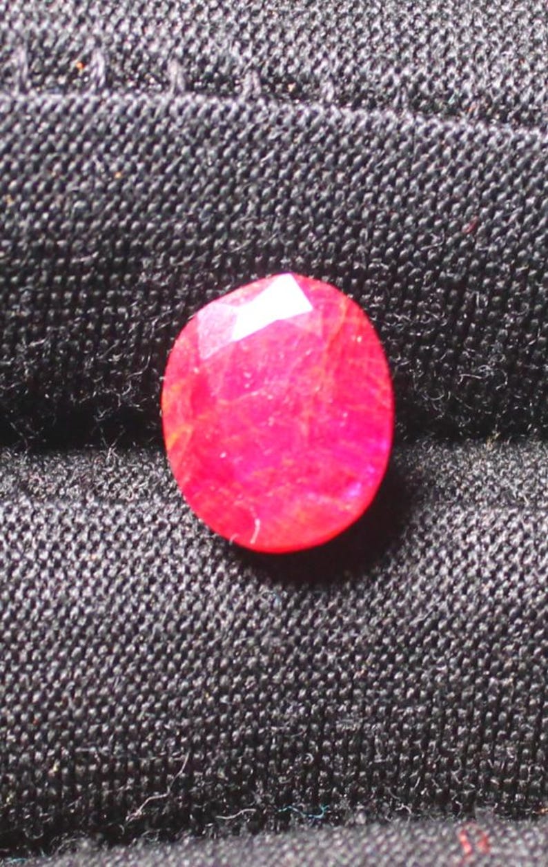 Puede incluir: Una gema ovalada de color rojo vibrante con un corte facetado. La gema tiene una apariencia ligeramente transl&uacute;cida con inclusiones visibles.