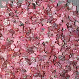 May include: A pile of small, rough, red ruby gemstones. The rubies are various shades of red and pink, and some have a slight sparkle.