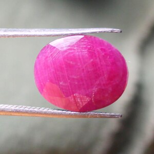 K&ouml;nnte beinhalten: Ein leuchtend rosa Rubin-Edelstein mit facettiertem Schliff, gehalten in einer Pinzette.