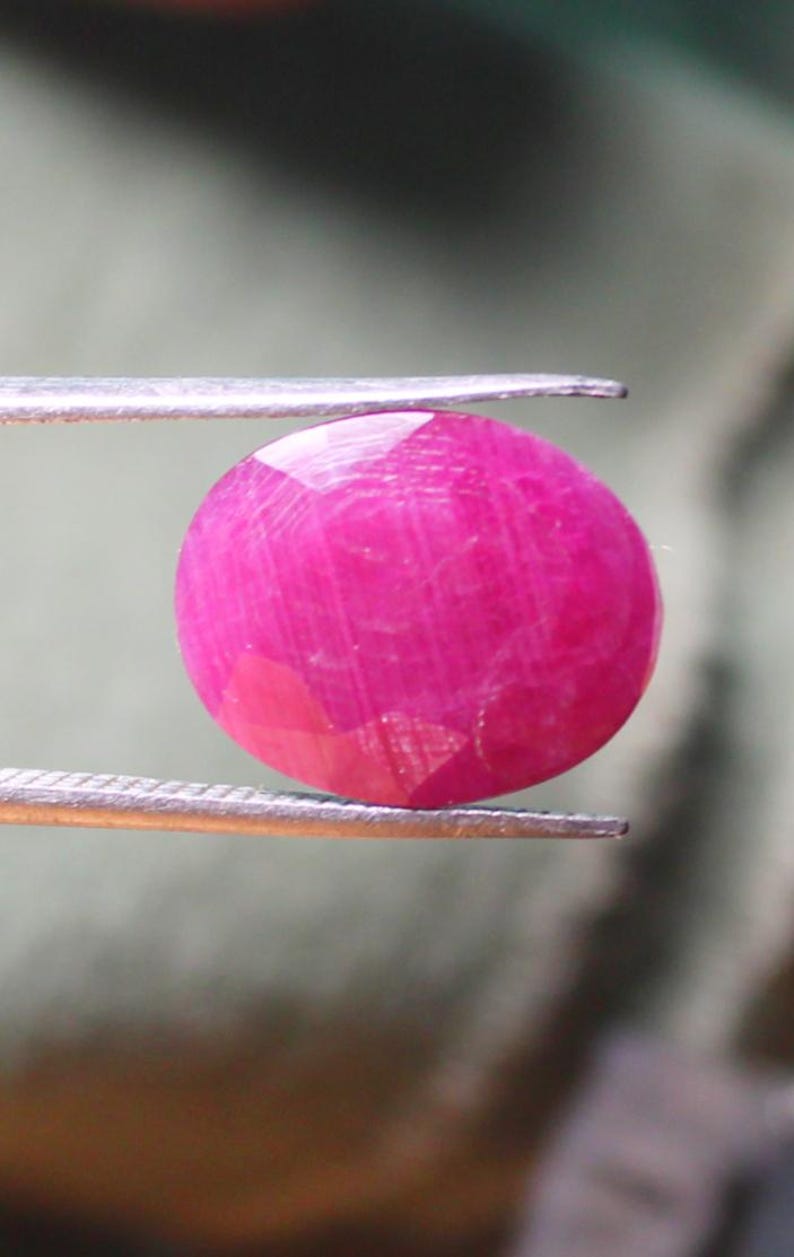 K&ouml;nnte beinhalten: Ein leuchtend rosa Rubin-Edelstein mit facettiertem Schliff, gehalten in einer Pinzette.