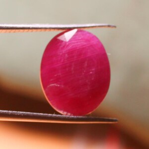 K&ouml;nnte beinhalten: Ein leuchtend rosa ovaler Edelstein, der mit einer Pinzette gehalten wird. Der Edelstein hat eine glatte, polierte Oberfl&auml;che und einen leichten Schimmer.