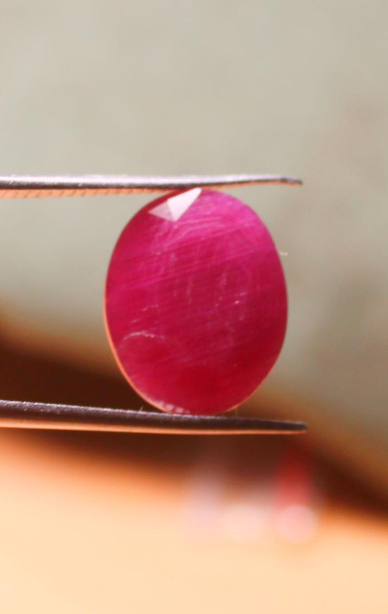 K&ouml;nnte beinhalten: Ein leuchtend rosa ovaler Edelstein, der mit einer Pinzette gehalten wird. Der Edelstein hat eine glatte, polierte Oberfl&auml;che und einen leichten Schimmer.