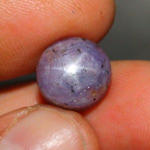 May include: A round, lavender-colored gemstone with a star-like pattern across its surface. The gem has a polished appearance and is held between two fingers, showcasing its spherical shape and subtle color variations. Small dark inclusions are visible.