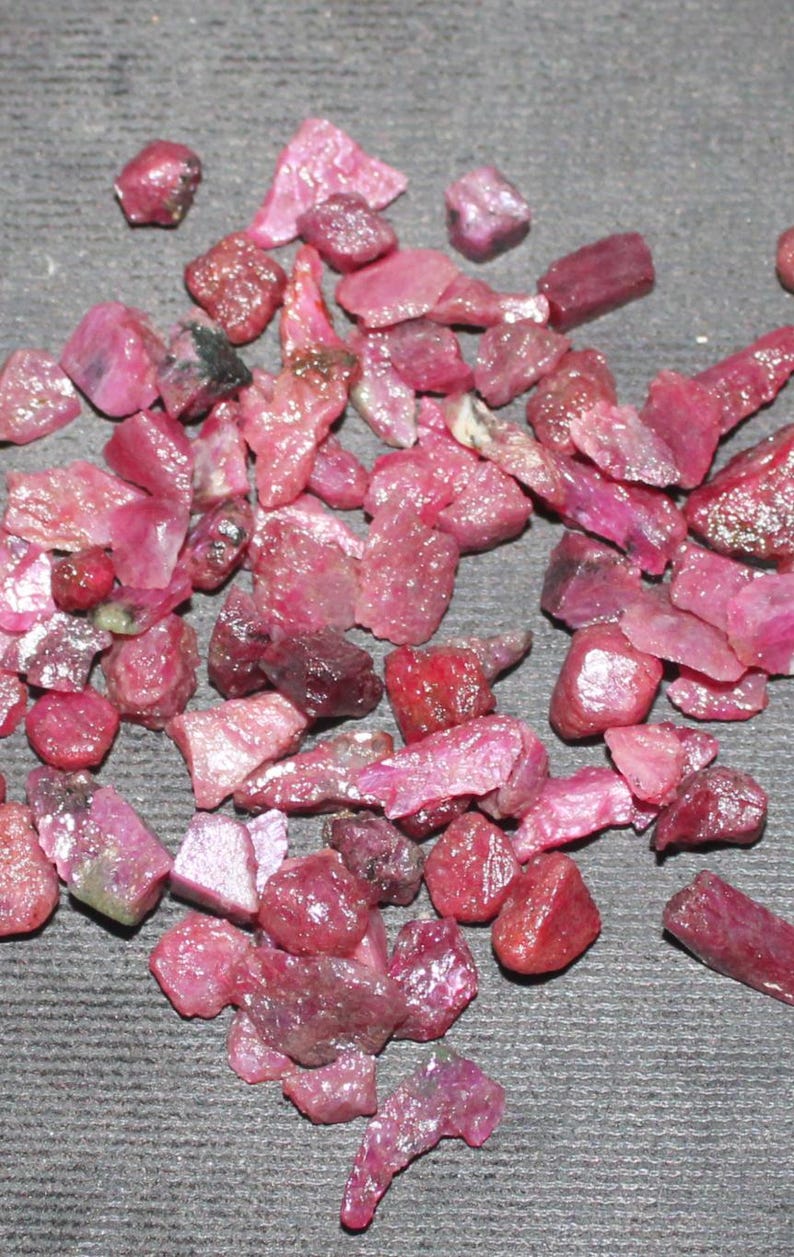 May include: A collection of raw, uncut ruby gemstones in varying shapes and sizes. The gemstones display a range of pink and red hues, with some having a deep, saturated color. The rough texture and natural forms of the rubies are visible.