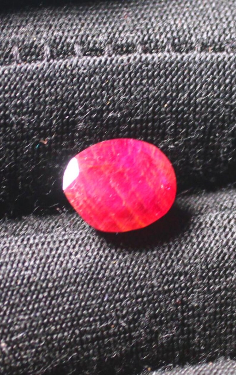 Puede incluir: Una piedra preciosa ovalada de color rojo intenso con una textura ligeramente rugosa.