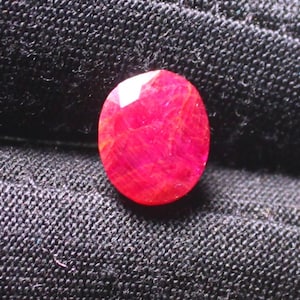 Puede incluir: Una piedra preciosa ovalada de color rojo oscuro con un corte facetado. La piedra preciosa est&aacute; colocada sobre un fondo de tela negra.