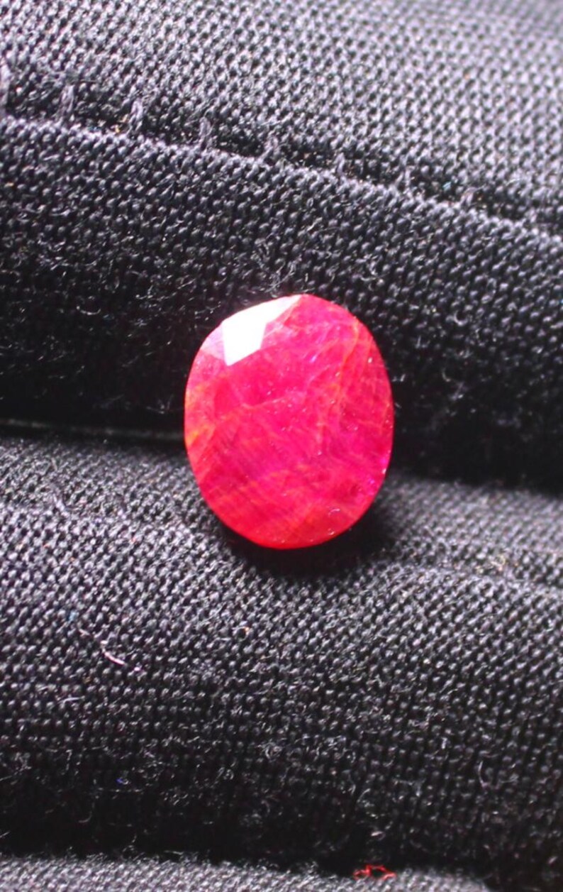 Puede incluir: Una piedra preciosa ovalada de color rojo oscuro con un corte facetado. La piedra preciosa est&aacute; colocada sobre un fondo de tela negra.