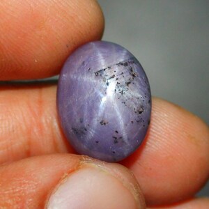 Op de afbeelding: Een paarse ster-saffier edelsteen met een zespuntige ster in het midden. De edelsteen is ovaalvormig en heeft een glad, gepolijst oppervlak.