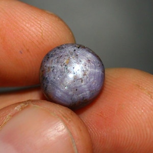 Peut inclure: Une pierre précieuse ronde de couleur violette avec un motif en forme d'étoile sur sa surface. La pierre précieuse est tenue dans une main.