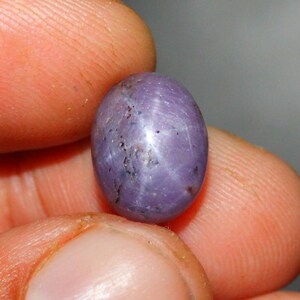 Puede incluir: Una piedra preciosa de zafiro estrella cabujón de color púrpura con una estrella de seis puntas.