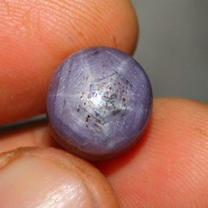 Op de afbeelding: Een ronde, paarse edelsteen met een zespuntige ster in het midden. De ster is zichtbaar door de manier waarop het licht van de steen reflecteert.