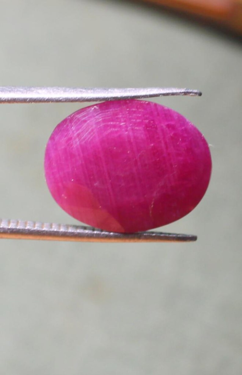 K&ouml;nnte beinhalten: Ein leuchtend rosa ovaler Edelstein, wahrscheinlich ein Rubin, wird von einer Pinzette gehalten.