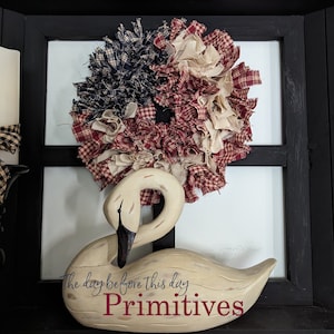 May include: A white wooden swan figurine with the text "The day before this day Primitives" on its base. A red, white and blue rag wreath is behind the swan.