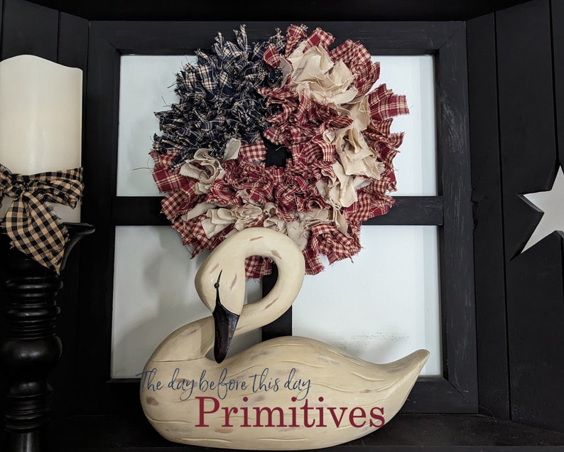 May include: A white wooden swan figurine with the text "The day before this day Primitives" on its base. A red, white and blue rag wreath is behind the swan.